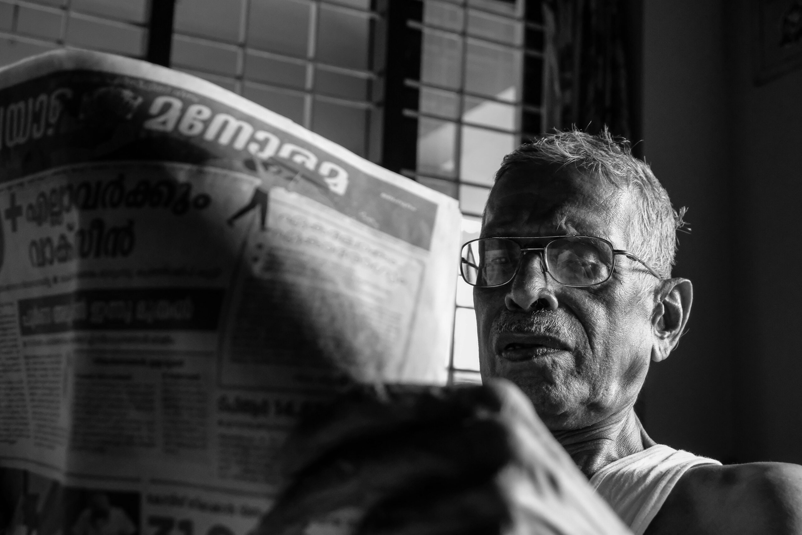 Black and white photo of an elderly man with glasses reading a newspaper in Nilambur, India.